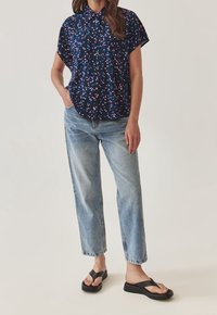 Marineblauw shirt met korte mouwen en kleurrijk abstract patroon, losse pasvorm; gecombineerd met lichtblauwe rechte jeans en zwarte flip-flops.
