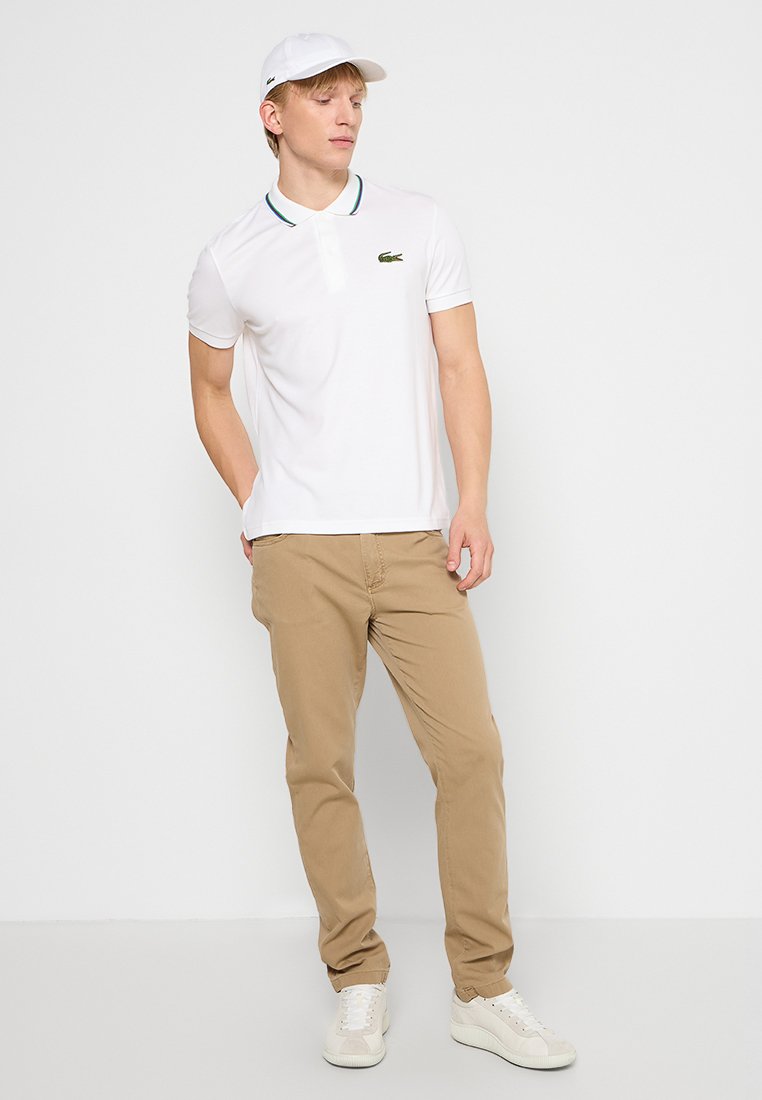 Polo shirt blanc avec un collier et des bordures vert-bleu, associé à un pantalon beige. Une casquette blanche et des baskets blanches complètent le look.