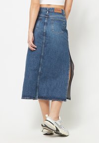 Femme portant une jupe midi en jean bleue avec des poches arrière et une fente latérale, associée à des baskets blanches de sport sur un fond uni.