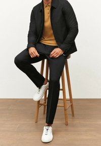 Sort jakke over en sennepsgul polo, kombineret med sorte bukser og hvide sneakers. Glat stof og tætsiddende design, siddende på en træskammel.