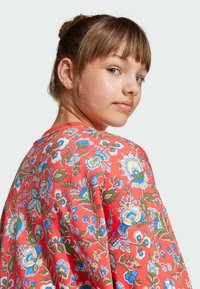 Halenka s kvetinovým vzorom v koralovej farbe s krátkymi rukávmi. Má modré a zelené kvety s prepracovanými detailmi a uvoľnený strih.