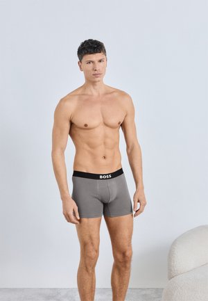 Graue Boxershorts mit einem schwarzen Bund, auf dem der Markenname "BOSS" deutlich zu sehen ist. Glatte Textur und körperbetonte Passform.