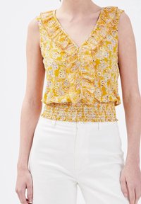 Haut jaune sans manches avec motif floral, col à volants et taille smockée. Porté avec un pantalon blanc, mettant en valeur les couleurs vives et les détails du design.