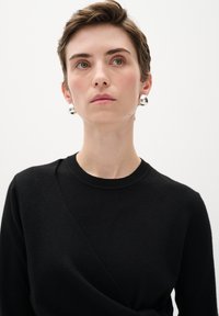Schwarzer Pullover mit rundem Ausschnitt und diagonalen Designelementen. Silberne Creolen verleihen dem Gesamtlook Glanz. Glatte Textur.