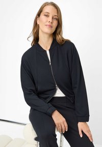 Schwarze Jacke mit Reißverschluss, rundem Ausschnitt, langen Ärmeln und lockerem Sitz. Das Material wirkt glatt, mit einem dezenten Glanz und ohne Muster.