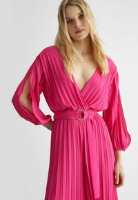 Abito plissettato di un vivace rosa con scollatura a V, maniche lunghe e vita con cintura dotata di una fibbia dorata. Texture morbida, design fluido.