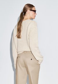 Pull en tricot beige avec un ourlet côtelé, associé à un pantalon beige clair doté de poches arrière. Cheveux coiffés en une queue de cheval basse.