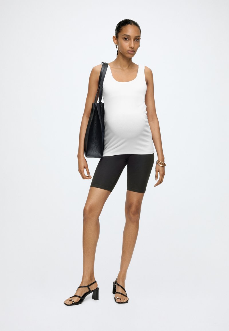 Gravid kvinde står iført en hvid tanktop, sorte cykelshorts, sorte hælsandaler og bærer en sort mulepose på venstre skulder.