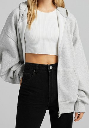 Femme portant un crop top blanc côtelé, un sweat à capuche zippé gris clair oversize, et un jean noir taille haute sur un fond uni.