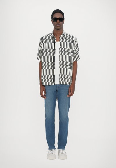 Chemise à manches courtes avec des rayures verticales noires et blanches, associée à un jean bleu clair et des baskets blanches. Le modèle porte des lunettes de soleil.