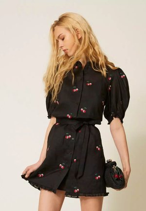 Mujer con cabello largo y rubio, que lleva una blusa negra con estampado de cerezas y una falda a juego, sosteniendo un pequeño bolso negro con decoración de cerezas.