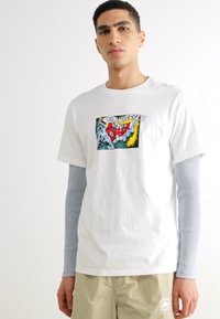 Camiseta blanca de algodón con un gráfico colorido que presenta manos y texto. Mangas largas y acanaladas de color azul claro visibles por debajo.