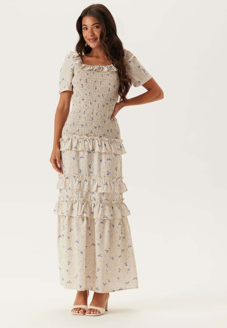 Blommig maxiklänning i ljusbeige med volangsteg, smockad liv, fyrkantig halsringning och korta puffärmar. Försköndad med små blå blommor.