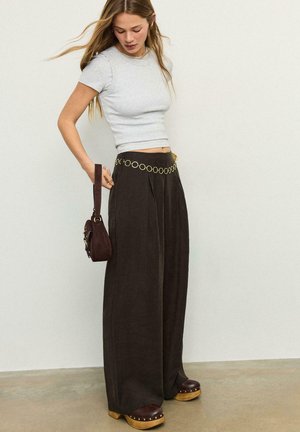 Kvinde iført en lysegrå cropped t-shirt, vidbenede mørkebrune bukser med guld cirkelformet kædebælte, bærende en mørkebrunt håndtaske, kiggende ned.