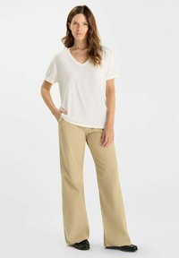 Vrouw met lang bruin haar, gekleed in een witte V-hals t-shirt, beige wijde broek en zwarte schoenen, staand met één hand in de zak.