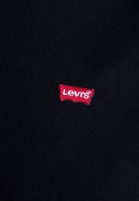 Etichetta con logo Levi's in rosso su tessuto blu scuro, con una texture liscia e tessuta. Il logo è ricamato con bordi frastagliati.
