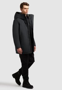 Sort isoleret parka med en stor hætte, frontknap og lynlåslukning, sidelommer og en glat ydre overflade. Båret med mørke cargo bukser og støvler.