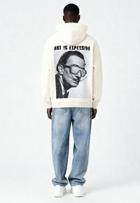 Hoodie crème avec un grand motif en noir et blanc d'un homme portant des lunettes et le texte "L'ART EST CHER." Porté avec un jean baggy bleu clair et des baskets blanches.