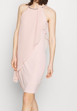 Frau trägt ein ärmelloses, blassrosa knielanges Kleid mit asymmetrischem Rüschensaum und verziertem Neckholder-Ausschnitt.