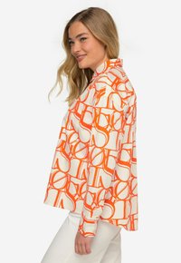 Camicia bianca con motivo circolare arancione, maniche lunghe e colletto. Il tessuto appare morbido, con una vestibilità comoda.