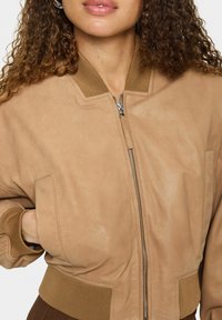 Frau trägt eine beige Wildleder-Bomberjacke mit geripptem Kragen, Bündchen, Saum und Frontreißverschluss, Hand in der linken Tasche, lockige Haare sichtbar.