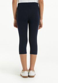 Námořnické legíny se strečovým materiálem a přiléhavým střihem, zkrácené nad lýtkem, spárované s bílými botami s hnědými podrážkami.