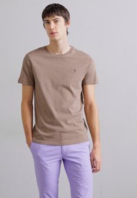 T-shirt justo castanho com decote redondo e pequeno logótipo no peito, combinado com calças lilás claras. Tecido suave, estilo casual.