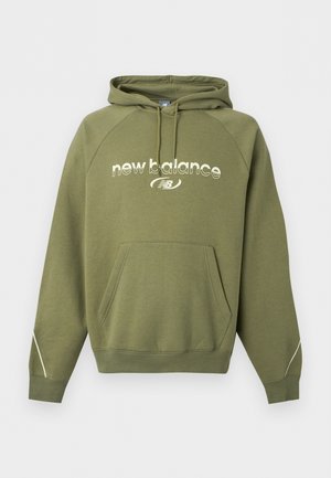 Jeune homme aux cheveux courts et bouclés portant un sweat à capuche New Balance vert olive avec une poche avant et un pantalon noir, debout sur un fond blanc.