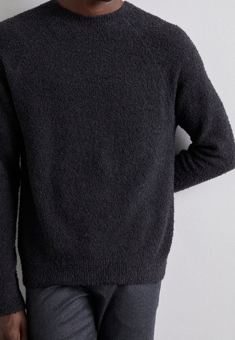 Schwarzer strukturierter Pullover mit Rundhalsausschnitt und langen Ärmeln, der aus einem weichen, flauschigen Material besteht und einen lässigen Schnitt hat, kombiniert mit dunkel gefärbten Hosen.