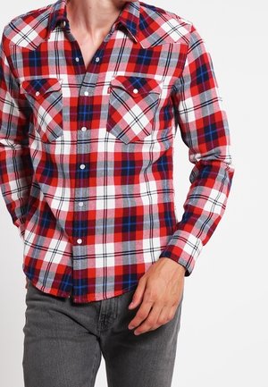 Homme portant une chemise à carreaux rouge, blanche et bleue boutonnée avec deux poches poitrine et un pantalon gris foncé sur fond blanc uni.