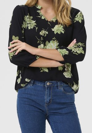 Woman wearing black blouse with green floral print and blue jeans, arms crossed at waist, blonde hair visible above shoulders.