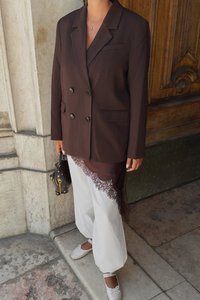 Brązowy żakiet dwurzędowy w połączeniu z białymi spodniami z akcentami z koronki. Nosi małą czarną torebkę. Całość dopełniają białe baleriny.