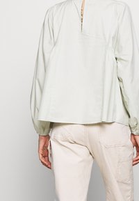 Person in einem hellbeigen Langarmshirt mit Rückenschlitz und off-white Hosen mit Gesäßtaschen, steht vor einem schlichten Hintergrund.
