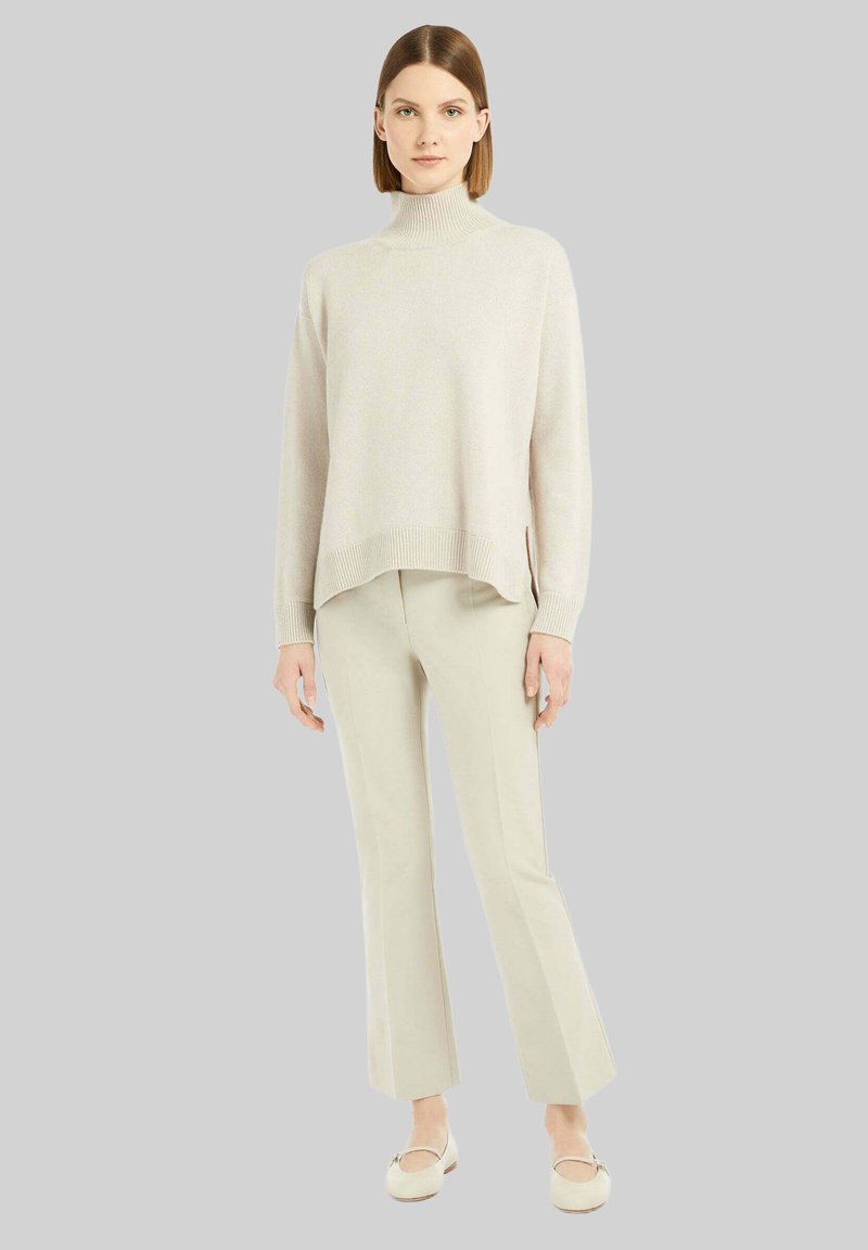 Maglione a collo alto beige chiaro con vestibilità comoda, fessure laterali e dettagli a costine, abbinato a pantaloni chiari e scarpe basse.
