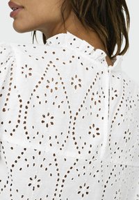 Weißes Baumwollkleidungsstück mit intricate Broderie Anglaise Muster und floralen Designs. Volant-Ausschnitt und Knopfverschluss hinten.