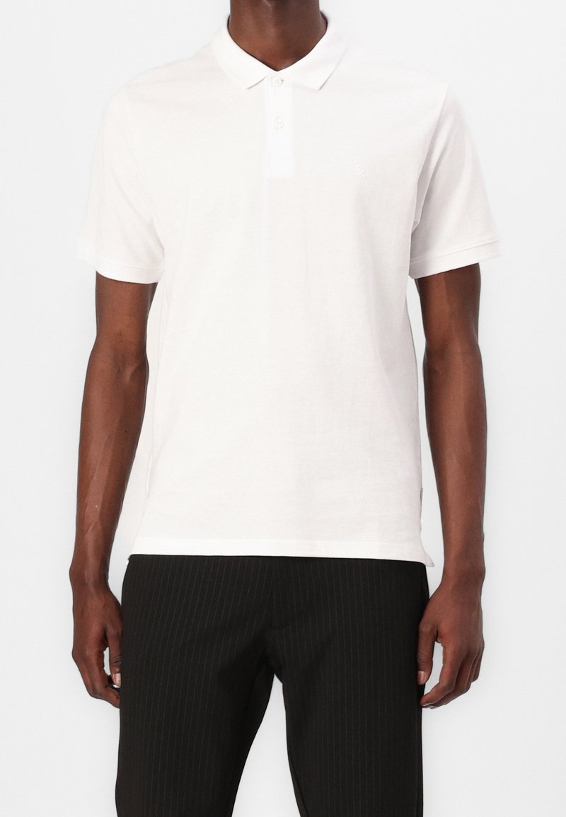 Homme portant un polo blanc uni à manches courtes et un pantalon noir à fines rayures, debout devant un fond blanc.