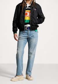 Svart bomberjacka, randig polo i flera färger, blå jeans och beige sneakers med röda och blå detaljer, med en texturerad tyg och synlig logotyp.