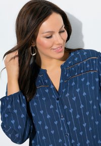 Donkerblauwe blouse met lichtblauw hartjespatroon, voorzien van een knoopsluiting aan de voorkant, geplooid ontwerp en elastische mouwen voor een zachte textuur.