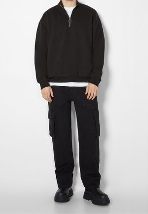 Felpa nera con zip e tirante ad anello, indossata sopra una t-shirt bianca. Abbinata a pantaloni cargo neri con tasche laterali e stivali neri.