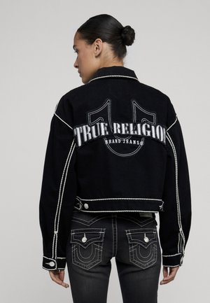 Frau mit dunklen Haaren im Dutt trägt eine schwarze True Religion-Jacke und schwarze Jeans mit kontrastierender weißer Naht, von hinten gesehen.
