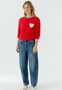 Rode zachte trui met een wit hartontwerp, ronde hals en driekwart mouwen, gecombineerd met een hoog getailleerde blauwe spijkerbroek.