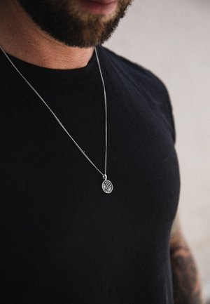 Sølv kæde halskæde med oval vedhæng, der viser et detaljeret design, båret over en sort t-shirt. Nærbillede af brystområdet.