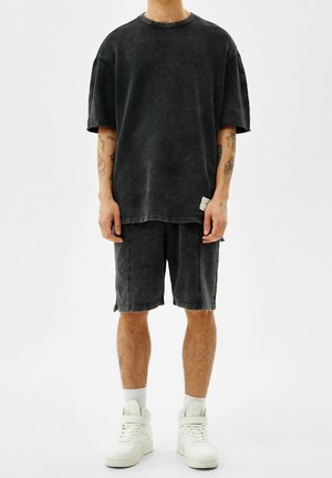 Person iført en falmet sort oversized T-shirt og matchende shorts, med hvide højtopede sneakers og synlige tatoveringer på armene, stående på hvid baggrund.