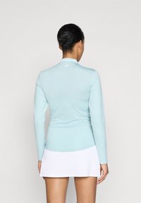 Camisa de manga longa azul clara com colarinho, textura suave e um pequeno logótipo branco nas costas, combinada com uma saia plissada branca.