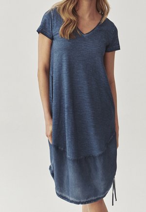 Woman wearing a short-sleeve navy blue casual dress with a v-neck and a slightly lighter denim-like hem, standing against a plain background.