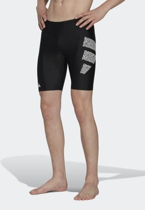 Mann trägt schwarze knielange Badeshorts mit drei weißen, gemusterten Streifen am rechten Oberschenkel, steht barfuß da und hat die Hände in die Hüften gestützt.