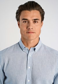 Giovane uomo con capelli scuri e occhi marroni che indossa una camicia azzurro chiaro con bottoni, guarda direttamente nella telecamera con un'espressione neutra.