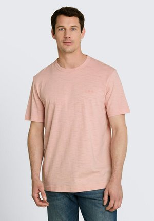 Man with short dark hair wearing a light pink short-sleeve T-shirt and blue jeans, standing against a plain light background.