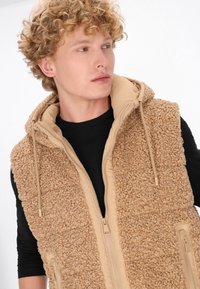 Gilet in pile color sabbia con un'esterno strutturato e peloso e un rivestimento interno liscio. Presenta una chiusura frontale con zip e un cappuccio regolabile con cordino. Camicia nera sotto.