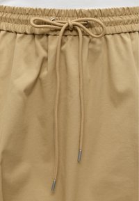 Ελαστική μέση με μπεζ κορδόνι δεμένο σε φιόγκο σε παντελόνι από ανοιχτό καφέ ύφασμα, που δείχνει την υφή και τα μεταλλικά τελειώματα στα άκρα του κορδονιού.